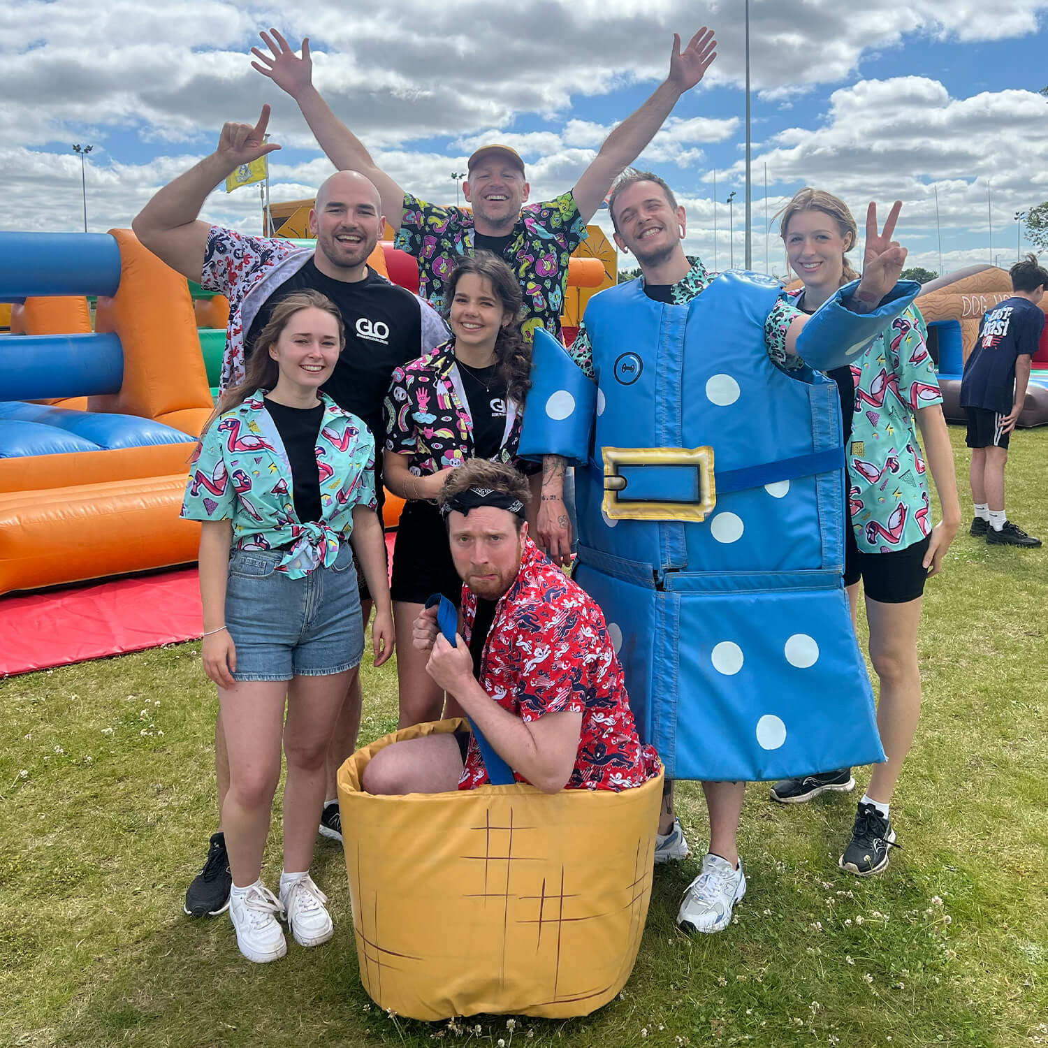 A group of seven people pose outdoors on grass, some wearing colorful shirts with fun patterns. One person is in a blue costume shaped like a vest, and another crouches in a basket costume. Inflatable obstacle courses are in the background.