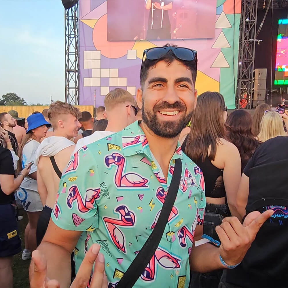 A man wearing a colorful flamingo shirt and sunglasses smiles and gestures with his hands at an outdoor music festival, surrounded by a crowd with a vibrant stage in the background.
