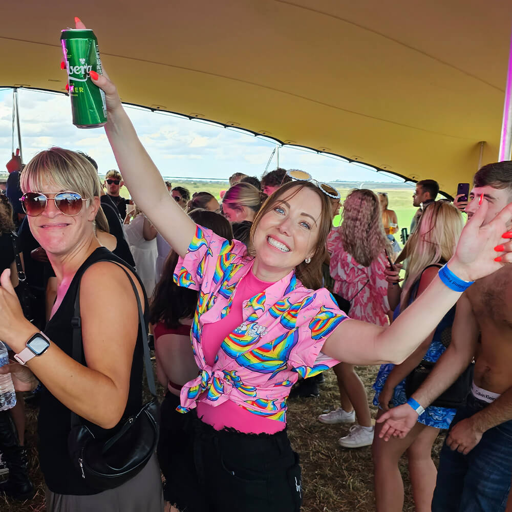 A smiling woman in a colorful shirt raises a can of beer and spreads her arms while dancing at an outdoor festival. People around her enjoy the event under a large tent with a sunny sky in the background.