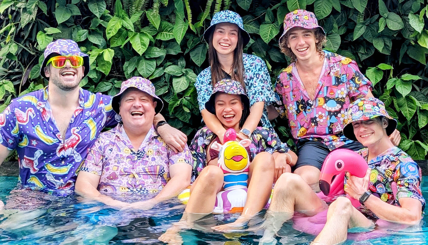 Six people wearing colorful shirts and bucket hats smile and pose together in a pool surrounded by green plants, with two inflatable toys—a duck and a flamingo—in front of them.