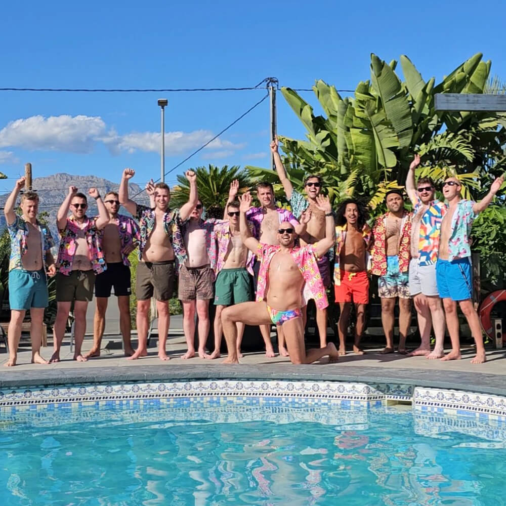A group of men in colorful shirts and swimwear stand and pose by a swimming pool on a sunny day, raising their arms and smiling, with lush green plants and a clear blue sky in the background.