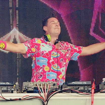 A DJ wearing the Badass Shirt, featuring a vibrant flamingo print, spreads his arms wide on stage with green headphones, DJ equipment, and bright LED lights in the background.