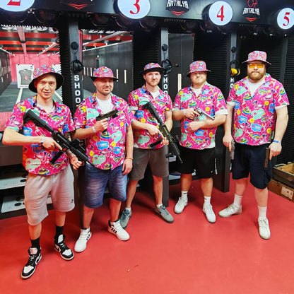 Five men at a shooting range pose with rifles, all wearing matching Badass Bucket Hats and colorful shirts that coordinate with the range’s red and black color scheme.