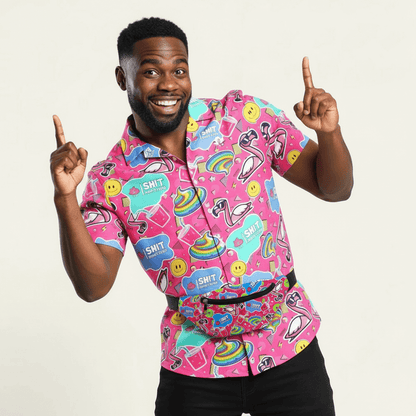 A smiling man points both index fingers up, wearing the Badass Shirt—a bright pink top adorned with rainbows, flamingos, smiley faces, and comic-style symbols—set against a plain light background.