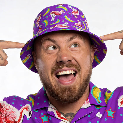 A bearded man smiles widely, pointing at his Bananas For Flamingos Bucket Hat, which complements his vibrant shirt covered in playful flamingo, star, rainbow, and bacon patterns.