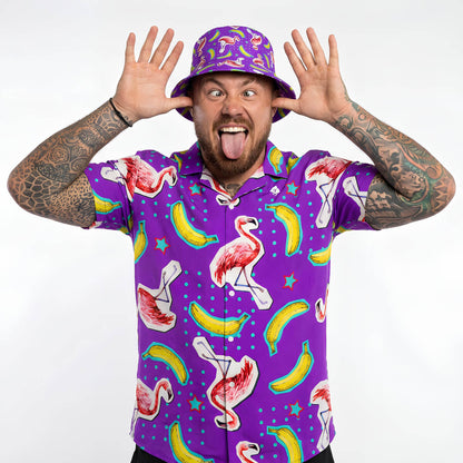 Wearing a bright purple Bananas For Flamingos Shirt, a person sticks out their tongue and holds their hands by their head in a playful pose against a plain white background.