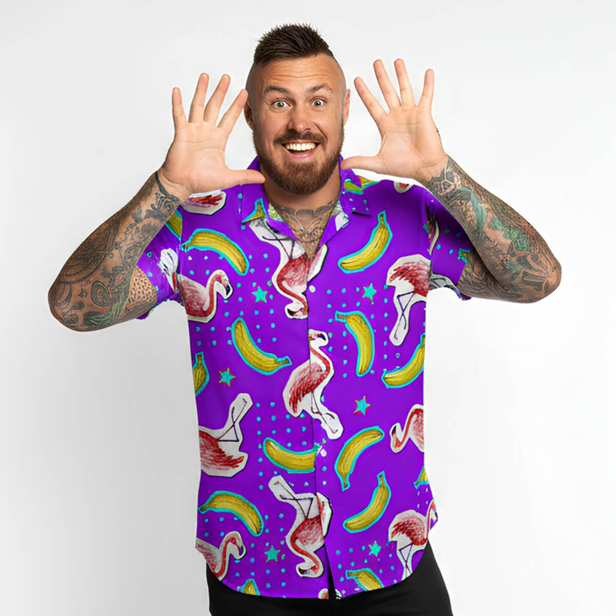 A smiling, tattooed man with a beard raises his hands to his face, wearing the Bananas For Flamingos Shirt—featuring colorful flamingos, bananas, and stars—while standing against a plain white background.