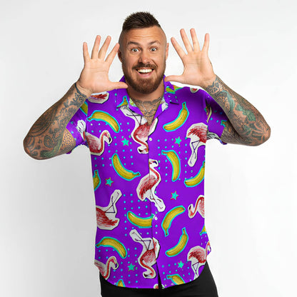 A smiling, tattooed man with a beard raises his hands to his face, wearing the Bananas For Flamingos Shirt—featuring colorful flamingos, bananas, and stars—while standing against a plain white background.