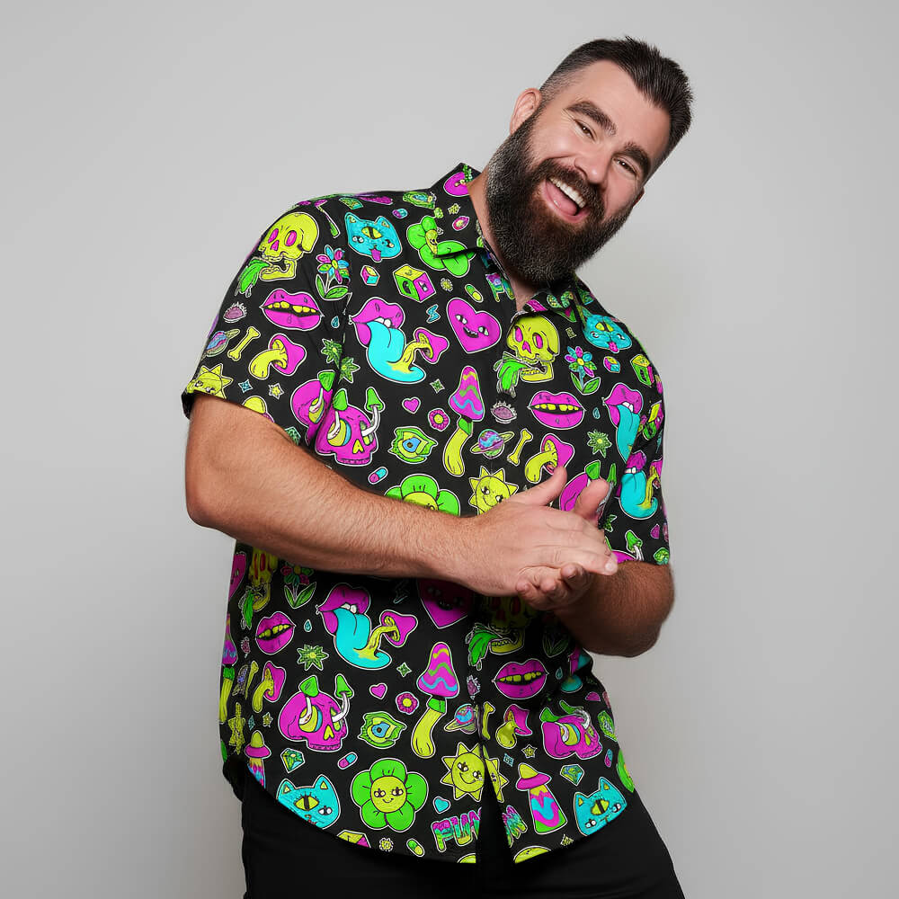 A bearded man with dark hair smiles and leans forward, wearing the Black Out Mushroom Shirt featuring neon cartoon faces, lips, frogs, and mushrooms—making a bold festival fashion statement against a plain light gray background.