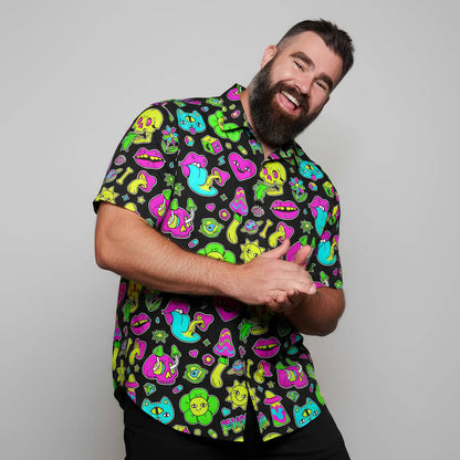 A bearded man with dark hair smiles and leans forward, wearing the Black Out Mushroom Shirt featuring neon cartoon faces, lips, frogs, and mushrooms—making a bold festival fashion statement against a plain light gray background.