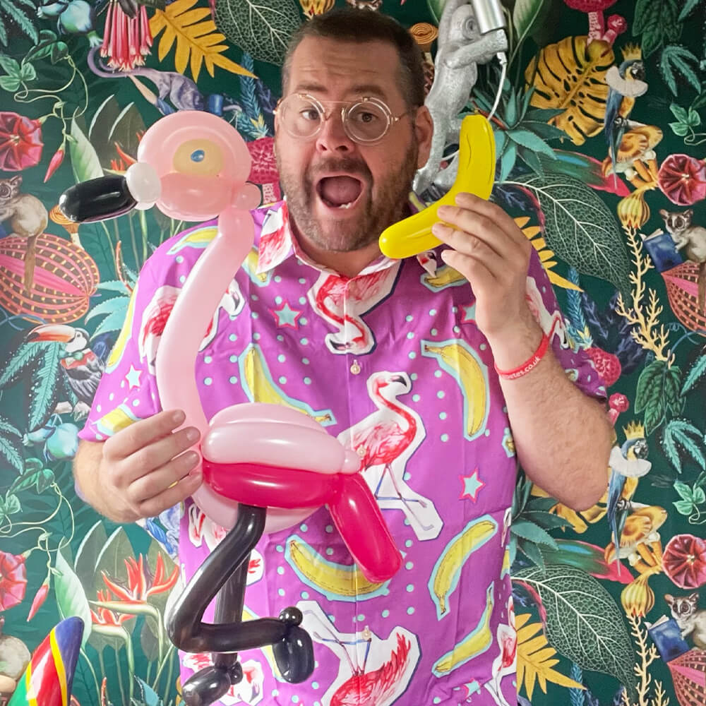 A man in a pink flamingo-patterned shirt holds a balloon flamingo and a yellow balloon shaped like a banana, standing in front of a colorful, tropical-patterned background.