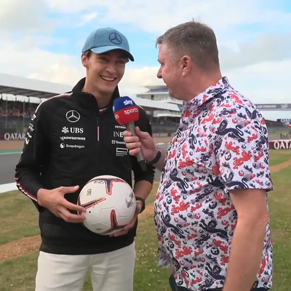 A smiling man in a Mercedes jacket and blue cap holds a soccer ball while being interviewed by another man in a colorful floral shirt holding a Sky Sports microphone at a racetrack.
