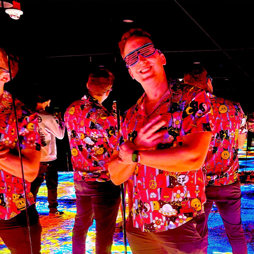 A man wearing a colorful, cartoon-patterned shirt and light-up shutter shades poses with his arms crossed in front of mirrored walls, creating multiple reflections. The floor is brightly lit with multicolored patterns.
