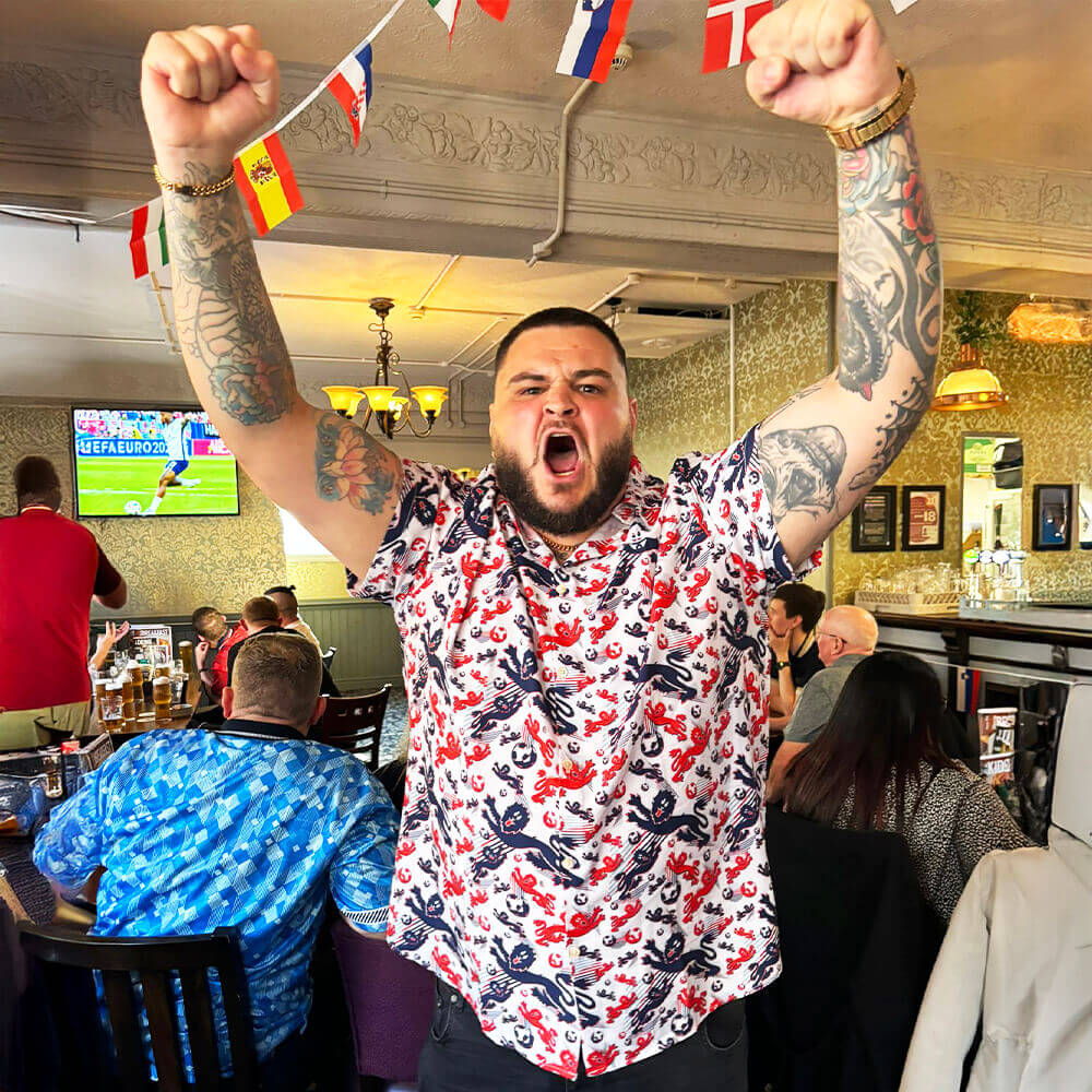A bearded man with tattooed arms, wearing the England Football Hawaiian Shirt, cheers with both fists raised in a lively pub adorned with international flags as others watch a soccer match on TV behind him.
