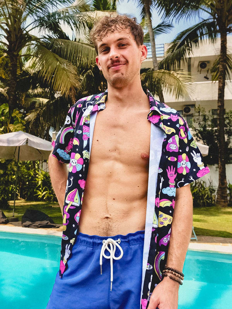 A young man with curly hair stands by a pool, wearing blue swim shorts and an open, colorful short-sleeve shirt with cartoon designs. Palm trees and a modern building are in the background.
