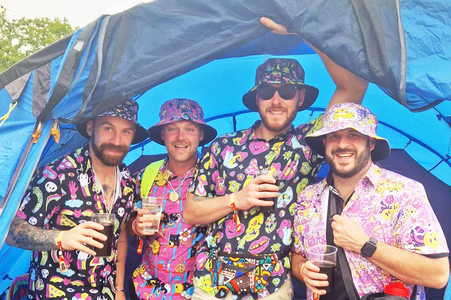 Four smiling men stand together in front of a blue tent, wearing colorful patterned shirts and matching bucket hats. They are each holding drinks and appear to be enjoying a festival or outdoor event.