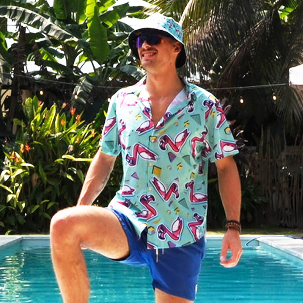 A man in the Flamingo Fandango Shirt, blue shorts, and a matching bucket hat stands by a pool with one leg raised and smiles, surrounded by lush tropical plants.