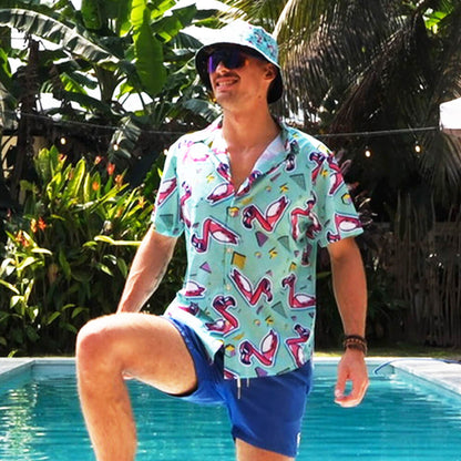 A man in the Flamingo Fandango Shirt, blue shorts, and a matching bucket hat stands by a pool with one leg raised and smiles, surrounded by lush tropical plants.