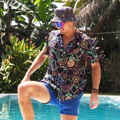 A man wearing a Fluorescent Fruit Fiesta Bucket Hat, a colorful fruit-patterned shirt, blue shorts, and reflective sunglasses stands by a pool with one knee lifted, surrounded by lush tropical plants.
