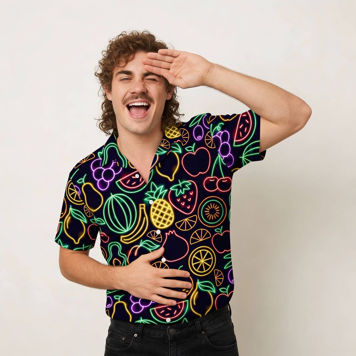 A cheerful man with curly hair and a mustache wears the Fluorescent Fruit Fiesta Shirt, featuring neon fruit patterns. He playfully salutes with one hand and rests the other on his stomach, posing against a plain background.