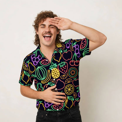 A cheerful man with curly hair and a mustache wears the Fluorescent Fruit Fiesta Shirt, featuring neon fruit patterns. He playfully salutes with one hand and rests the other on his stomach, posing against a plain background.