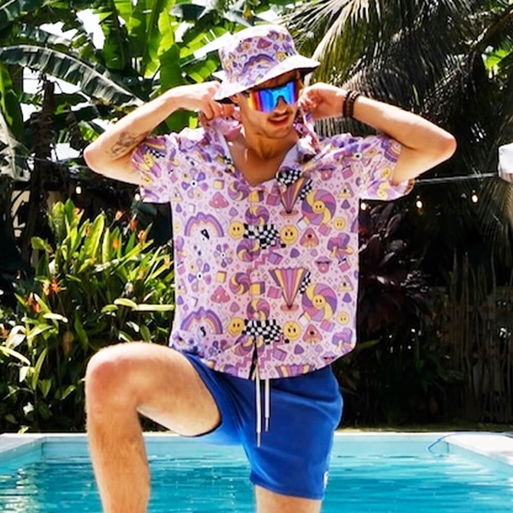 A person in a Hippie Bucket Hat, colorful sunglasses, pastel patterned shirt, and blue shorts poses playfully by a pool with tropical plants in the background.