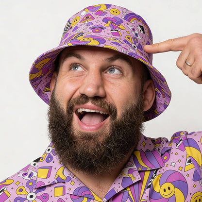 A bearded man grins as he points to his Hippie Bucket Hat, featuring playful abstract patterns in yellow, pink, and blue. His matching shirt shares the same vibrant psychedelic design.