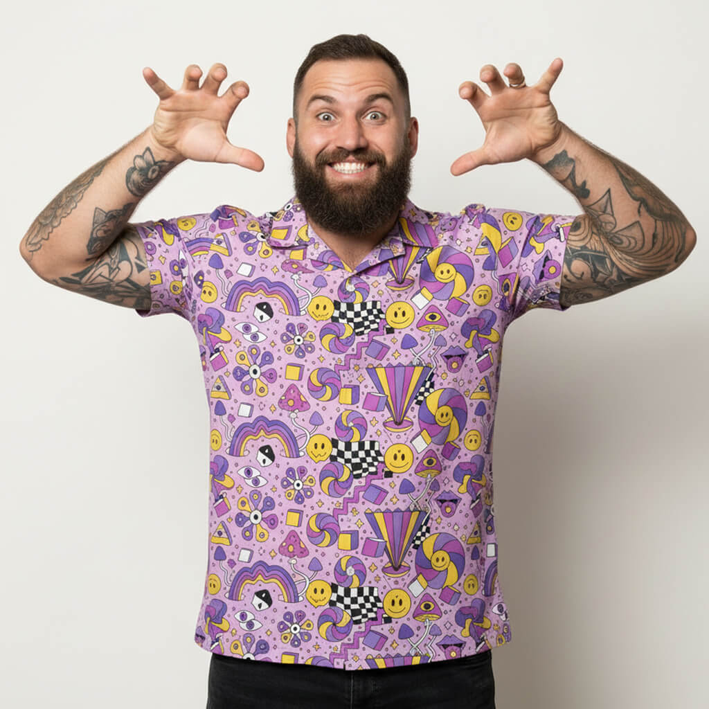 A bearded, tattooed man smiles widely and playfully raises his hands, showing off his palms while wearing a colorful Hippie Shirt with smiley faces and geometric patterns, standing against a plain white background.