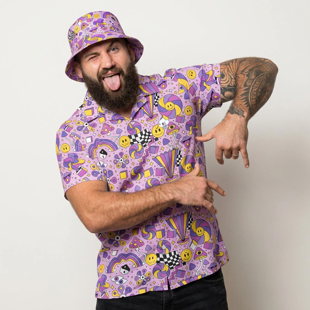 A bearded man playfully poses in the Hippie Shirt, featuring colorful 60s-inspired designs with smiley faces, stars, and abstract patterns. He sticks out his tongue, winks, and points at his hand while wearing a matching bucket hat.