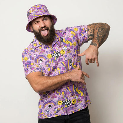 A bearded man playfully poses in the Hippie Shirt, featuring colorful 60s-inspired designs with smiley faces, stars, and abstract patterns. He sticks out his tongue, winks, and points at his hand while wearing a matching bucket hat.