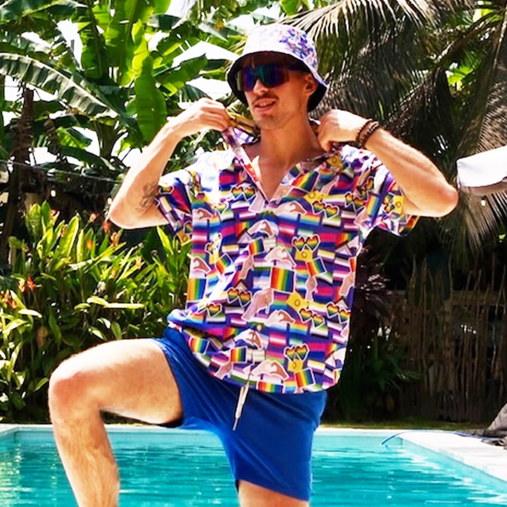 Wearing sunglasses and an LGBTQ Pride Bucket Hat, a person in a patterned shirt and blue shorts poses by a pool, lifting their collar. Lush green tropical plants fill the background.