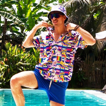 Wearing sunglasses and an LGBTQ Pride Bucket Hat, a person in a patterned shirt and blue shorts poses by a pool, lifting their collar. Lush green tropical plants fill the background.