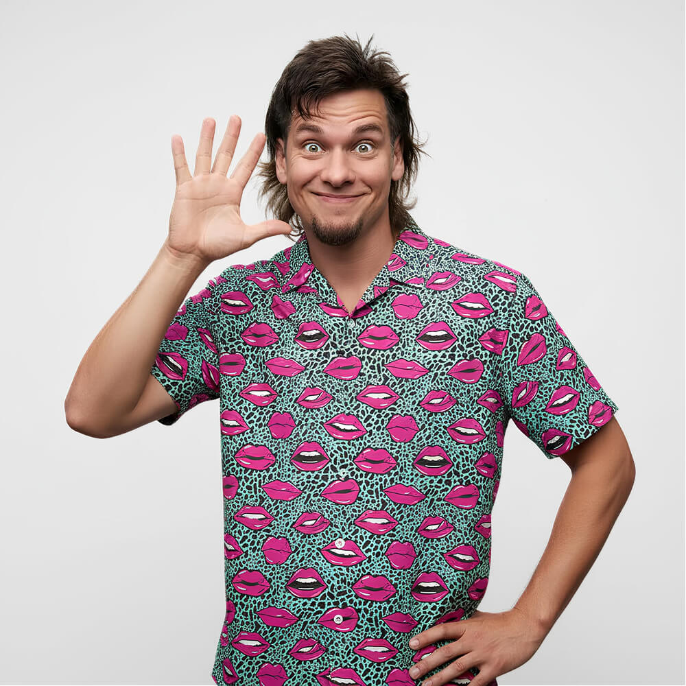 A smiling man with shaggy brown hair waves at the camera, wearing the Leopard Kiss Bad Shirt featuring pink and black cartoon lips, standing against a plain white background.