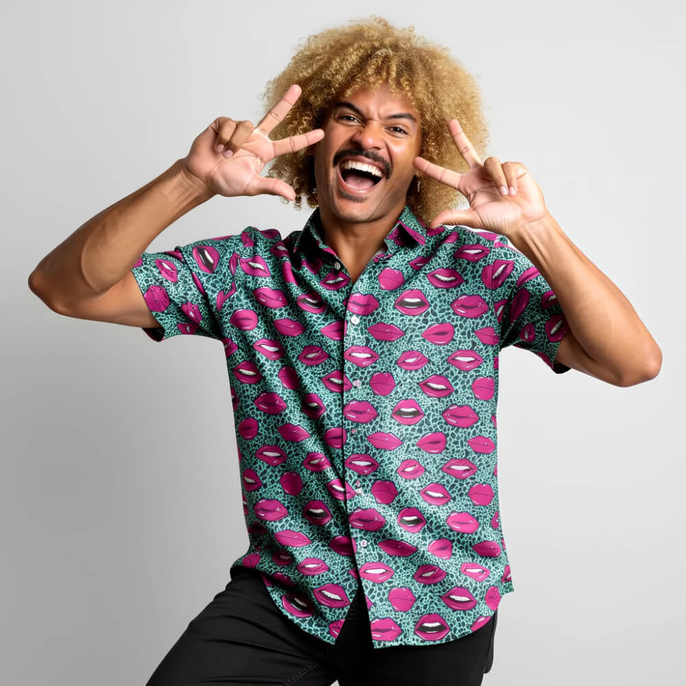 A smiling man with curly blonde hair makes peace signs, wearing the Leopard Kiss Bad Shirt featuring pink lips and geometric patterns, plus black pants, as he stands against a plain light gray background.