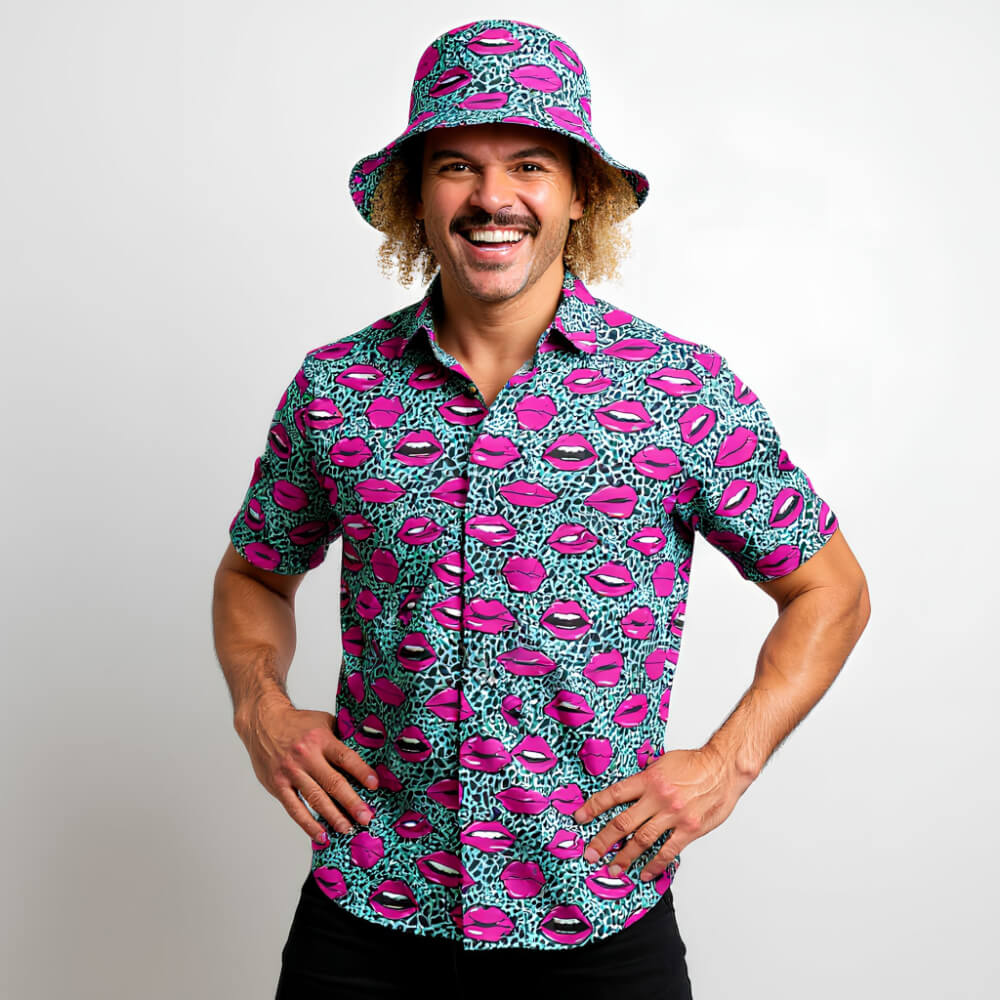 A smiling man with curly hair wears the Leopard Kiss Bad Shirt and matching bucket hat, both featuring pink lips and blue leopard prints, while standing against a plain white background.