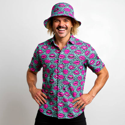 A smiling man with curly hair wears the Leopard Kiss Bad Shirt and matching bucket hat, both featuring pink lips and blue leopard prints, while standing against a plain white background.