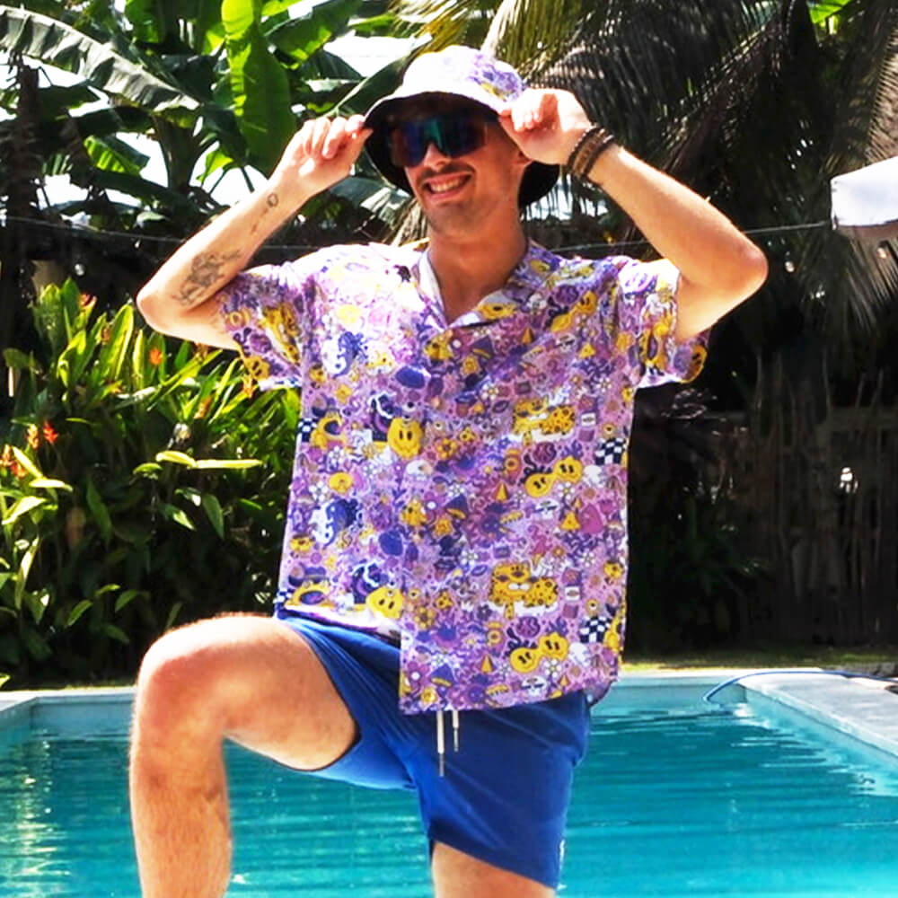 Wearing a vibrant purple shirt, blue shorts, and the Magical Psychedelic Bucket Hat, a person poses playfully by a pool amid tropical plants—showcasing the perfect festival-ready look.