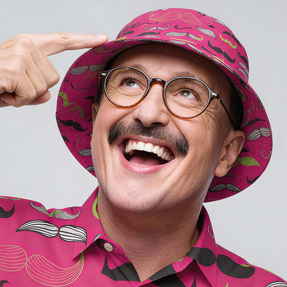 A smiling man with glasses and a mustache wears the Movember Bucket Hat and a mustache-patterned shirt. He points at his novelty hat and looks upward, standing against a light gray background.