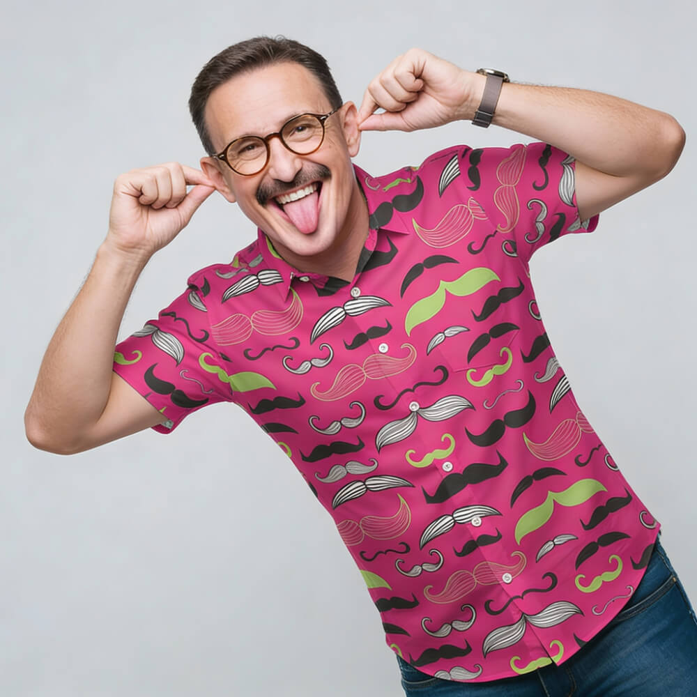 A man in glasses playfully pulls his ears and sticks out his tongue while wearing a bright pink Movember Shirt, featuring a mustache pattern, smiling against a plain light backdrop—showing his fun support for the Movember cause.