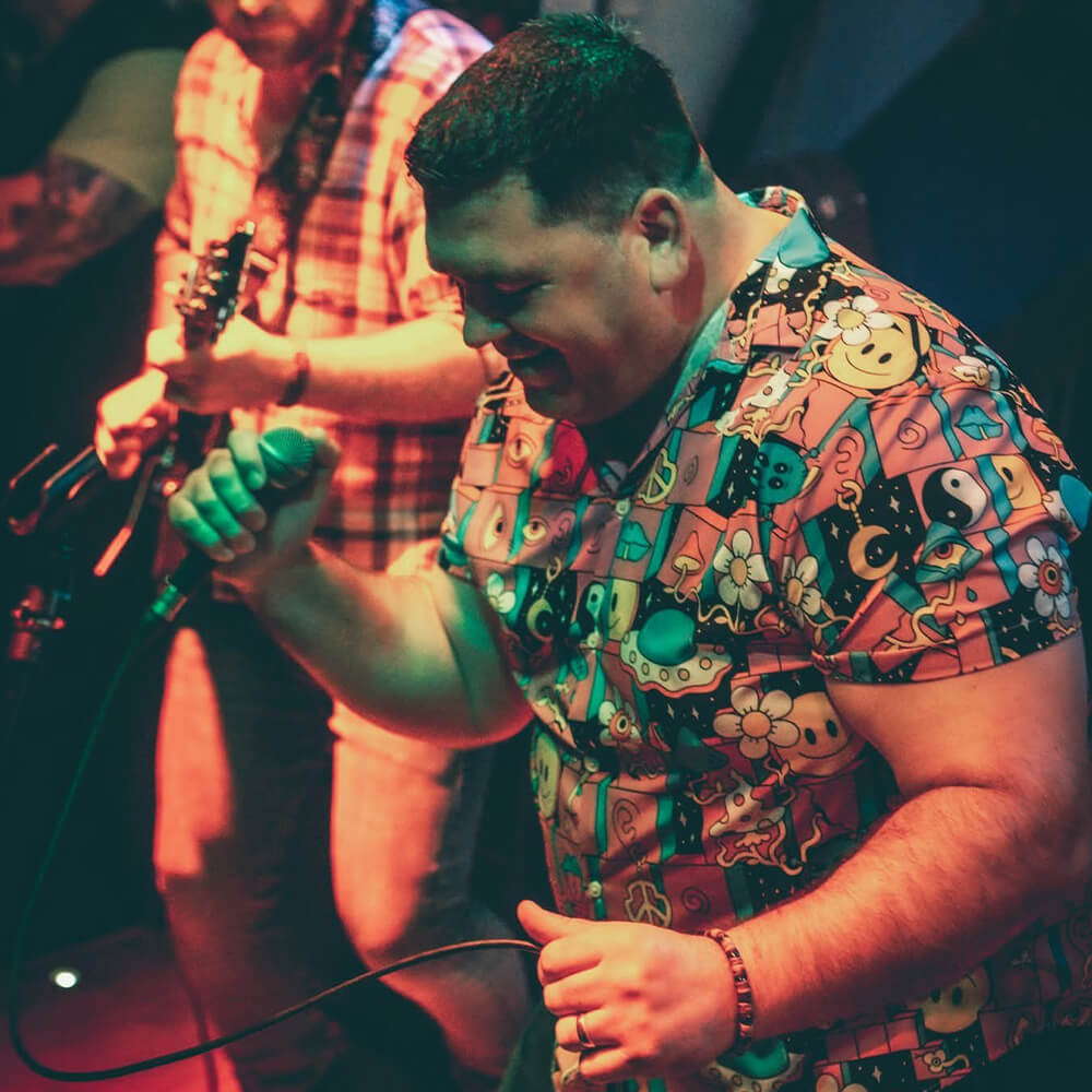 A man in a colorful, cartoon-patterned shirt passionately sings into a microphone on stage, while a guitarist plays in the background under vibrant lighting.