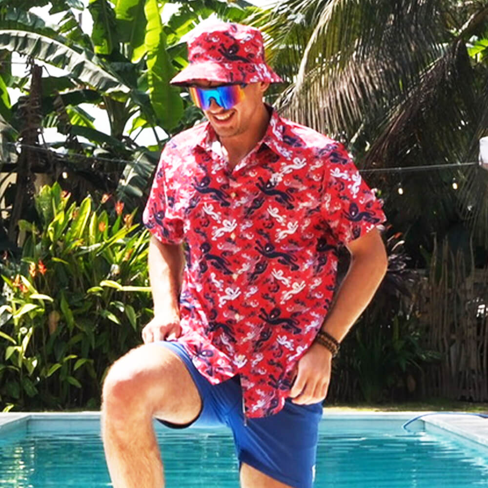 A smiling man shows off his English pride by the pool, sporting a Red England FC Hawaiian Shirt, matching bucket hat, blue shorts, and reflective sunglasses with tropical plants and palm trees in the background.