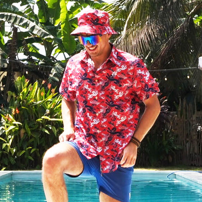 A smiling man shows off his English pride by the pool, sporting a Red England FC Hawaiian Shirt, matching bucket hat, blue shorts, and reflective sunglasses with tropical plants and palm trees in the background.