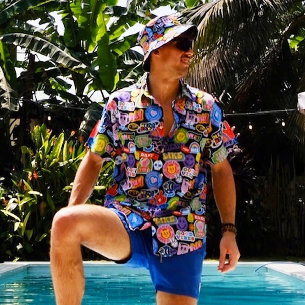 A man in a vibrant Retro Sticker Shirt and a matching bucket hat with smiley face designs stands poolside among lush greenery and palm trees, gazing off to the side with one leg raised.
