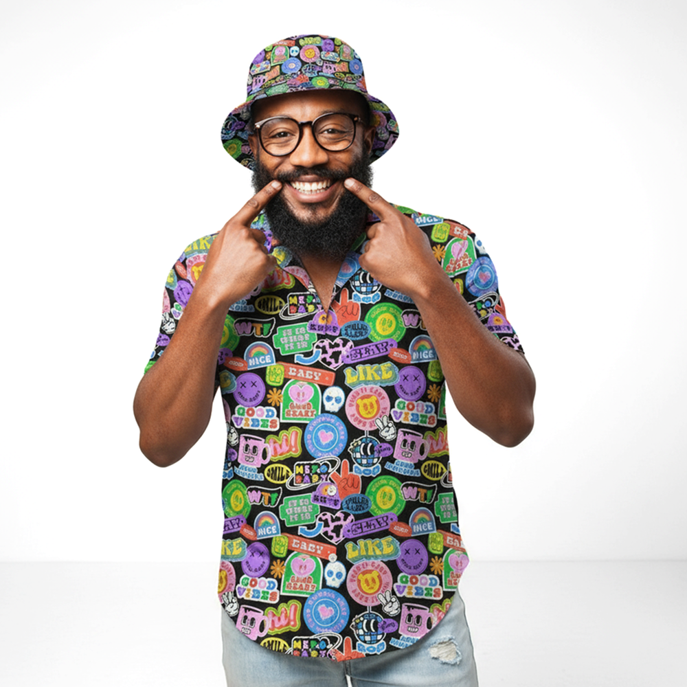 A bearded man with glasses wears the Retro Sticker Bucket Hat, featuring a colorful 90s pattern. He smiles, pointing to his grin with both index fingers while standing against a plain white background.