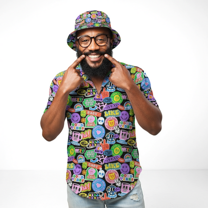 A bearded man with glasses wears the Retro Sticker Bucket Hat, featuring a colorful 90s pattern. He smiles, pointing to his grin with both index fingers while standing against a plain white background.