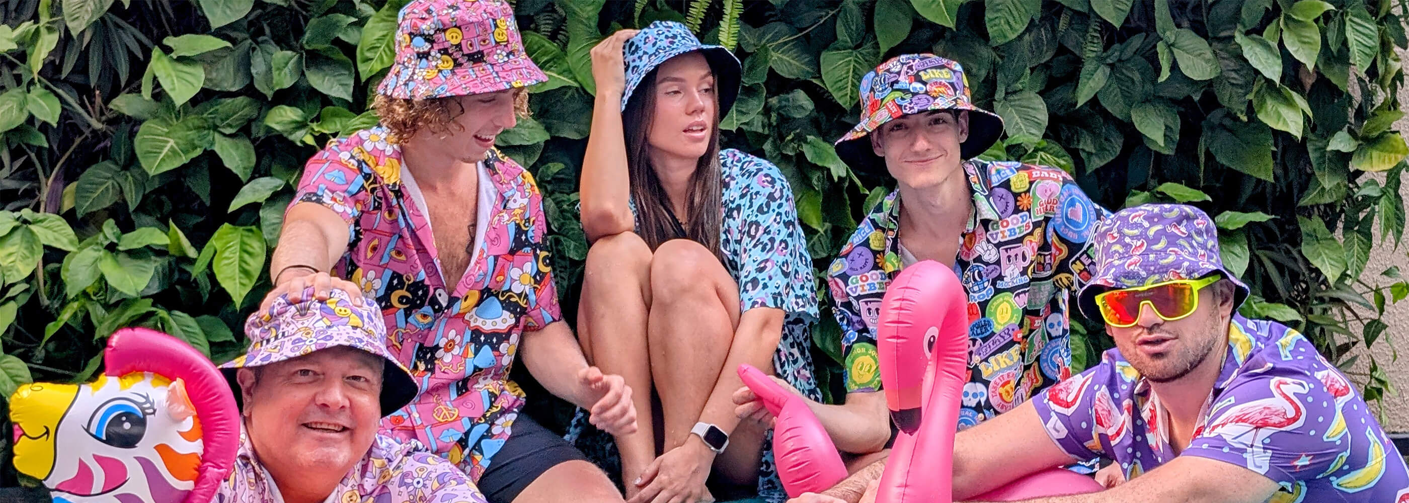 Five people in colorful, cartoon-patterned outfits and bucket hats sit together with inflatable toys, including a flamingo and unicorn, in front of lush green plants. They appear relaxed and playful.