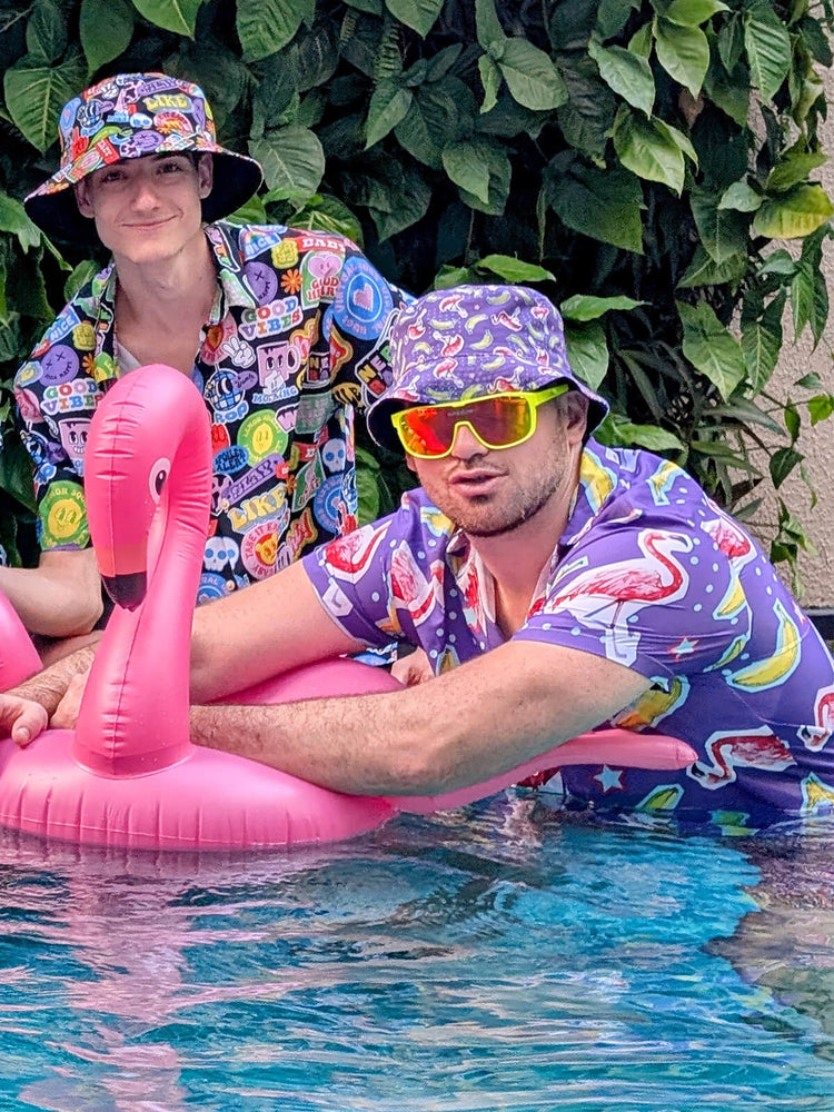 Two men in colorful shirts and bucket hats pose in a pool. One leans on a pink inflatable flamingo while wearing neon sunglasses. Green leaves are visible in the background.