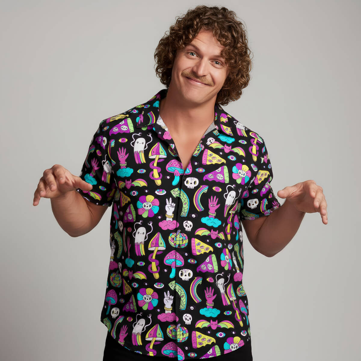 A man with curly hair and a mustache grins, playfully posing in the Shroom Shirt—featuring neon cartoon pizzas, skulls, mushrooms, and fun shapes—against a plain backdrop.