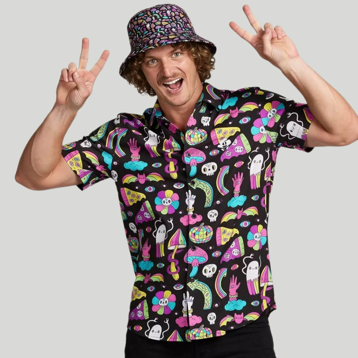 A smiling man in a colorful bucket hat and the Shroom Shirt, featuring vibrant neon mushroom and playful cartoon designs, makes peace signs with both hands against a plain light gray background.
