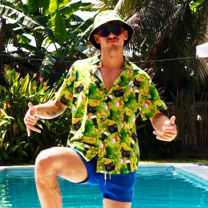 A man in sunglasses and a St Patrick’s Day Bucket Hat poses by a pool, wearing a bright green cartoon-print shirt and blue shorts, with tropical plants and palm trees in the background.